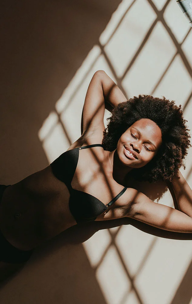 A woman with curly hair lying down with her eyes closed, illuminated by sunlight through a window frame.- Belt Lipectomy in Charlotte, NC