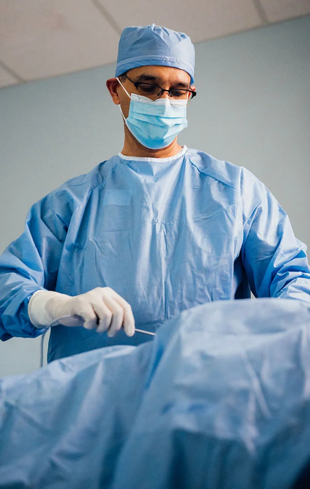 Dr. Haque, wearing blue surgical scrubs, a hair cap, and a face mask, focuses on performing a surgical procedure in an operating room environment. - Brazilian Butt Lift in Charlotte, NC