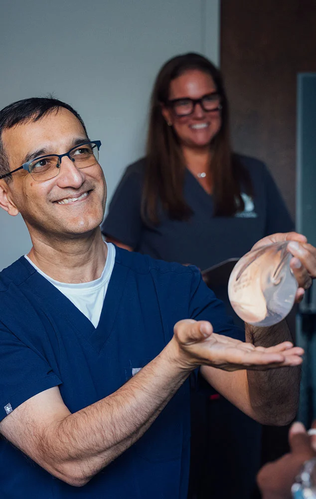 Dr. Haque smiling and holding up a breast implant in a clinical setting. - Breast Augmentation Revision in Charlotte, NC