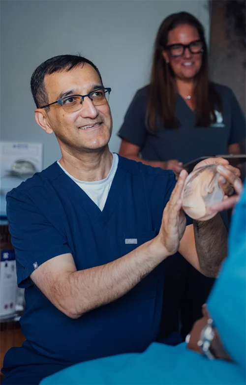Dr. Haque smiling and holding up a breast implant in a clinical setting. - Breast Augmentation in Charlotte, NC