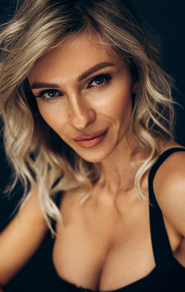Light-skinned woman with wavy blonde hair sits at a reflective table, leaning forward with hands gently placed together. She wears a dark sleeveless top and gazes directly at the viewer with a neutral expression. A solid black background contrasts with her light features. - Breast Lift with Augmentation in Charlotte, NC