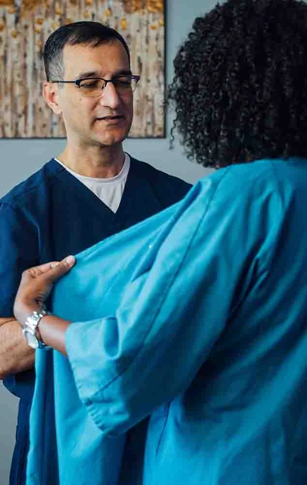 Dr. Haque wears dark blue scrubs and glasses while speaking with a patient wearing a teal medical gown. - Breast Reduction in Charlotte, NC