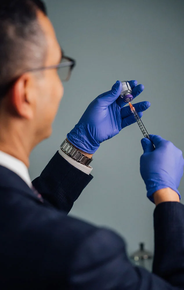 Dr. Haque wears blue sterile gloves while carefully drawing medication from a glass vial into a small syringe. - Dermal Fillers in Charlotte, NC