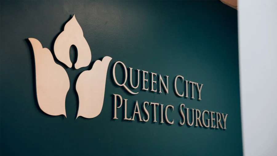 The Queen City Plastic Surgery logo mounted on a dark teal wall.