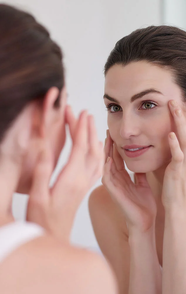 A young woman with long, dark hair looks intently at her reflection in a mirror, her face framed by her hands as she gently touches the skin around her eyes, possibly examining or applying a product. She is wearing a simple white tank top, and the background is a soft, blurred reflection, creating a sense of intimacy and focus on her skincare routine. - Lower Blepharoplasty in Charlotte, NC