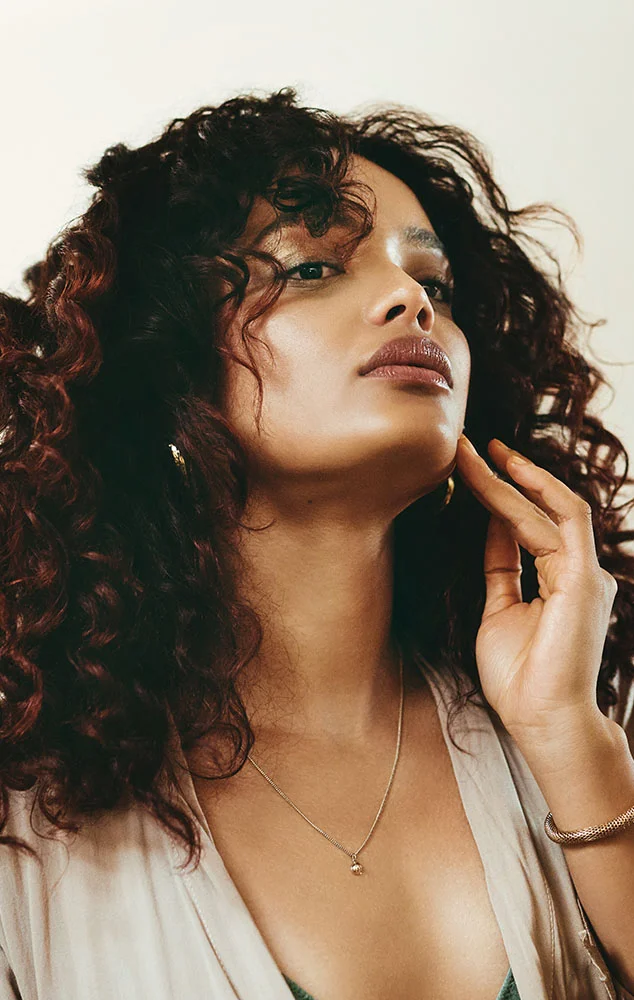 A young woman with long, curly brown hair, wearing a beige robe with lace trim. She is looking at her reflection in a mirror and touching her chin with her right hand. She is wearing a thin gold necklace and a beaded bracelet. - Neck Contouring in Charlotte, NC