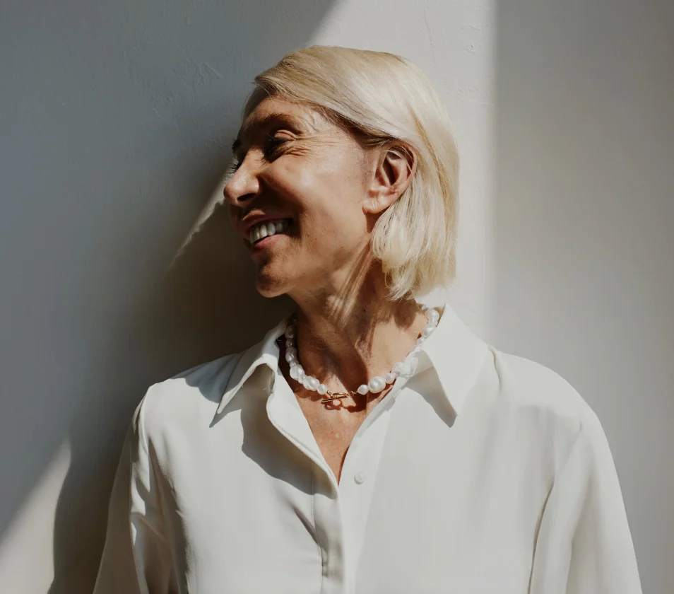 A woman with short blonde hair gazes upward and slightly to her right with a soft, cheerful expression. She wears a white button-down shirt and a pearl necklace with a small gold-toned pendant. The light gray background features distinct shadows that add depth, while the soft lighting highlights her face and creates a natural, portrait-like effect.