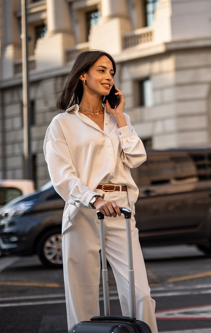 Woman in white clothing holding a suitcase and talking on a phone in a city.